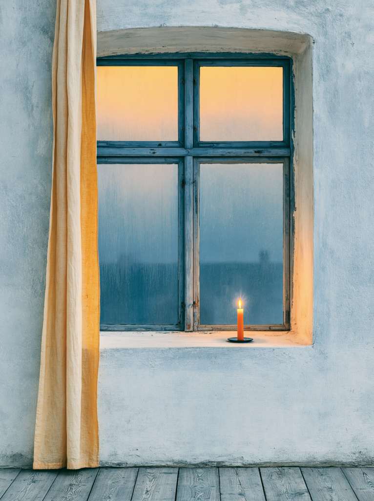A warm minimalist interior seen from inside on a dark winter evening a single candle burning on a bare windowsill, condensation on the cold glass beyond, the dark blue-black of night outside contrasting with the amber warmth within, pale oak floor, white walls, the window as a frame for the contrast between cold and comfort interior editorial photography, hygge atmosphere, deep navy and warm amber, cinematic and quiet